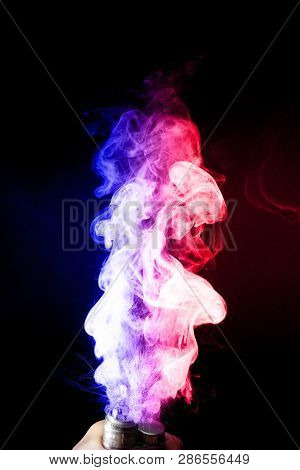Male Hand Holds Vape. Vape Clouds Splash From Vape Coil On Black Background. Fog Is Dual Color -- Bl