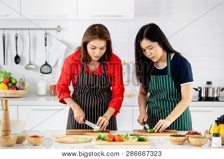 Beautiful Asian Woman In Red Shirt And Black  Apron Teaching Her Daughter How To Prepare Vegetables 