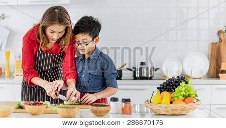 Asian Family Mom In Red Shirt And Black  Apron Is Teaching Her Son Use  Tool To Peel Fruit And Veget