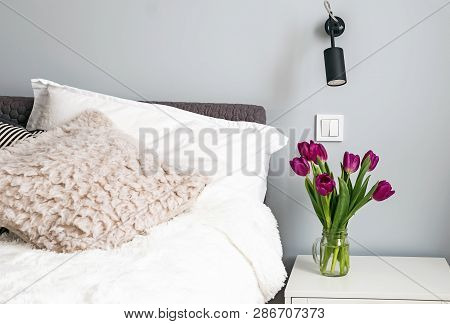 Purple Tulips Standing On A Nightstand In The Modern White Bedroom Near The Bed.