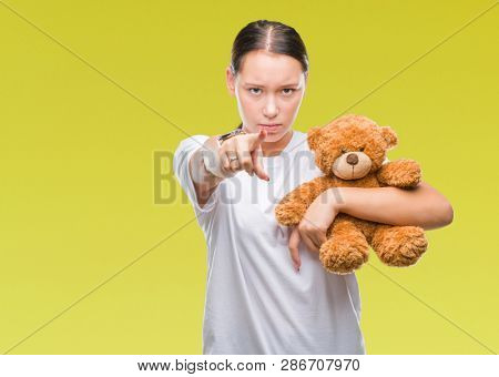 Young caucasian woman holding teddy bear over isolated background pointing with finger to the camera and to you, hand sign, positive and confident gesture from the front