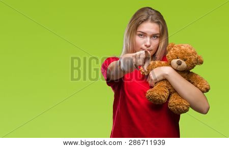 Young caucasian woman holding cute teddy bear over isolated background pointing with finger to the camera and to you, hand sign, positive and confident gesture from the front