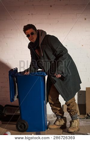 Homeless Man In Dark Glasses Rummaging In Blue Trash Container