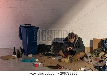Homeless Man Using Smartphone While Sitting On Cardboard Surrounded By Rubbish Dump