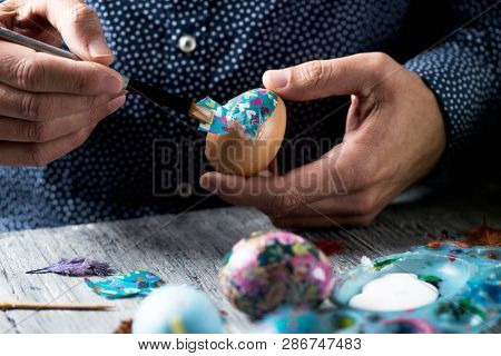 closeup of a young cacausian man decorating some homemade easter eggs by gluing some pieces of different papers, on a gray rustic wooden table