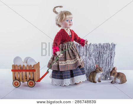 Cute Girl-doll With Trolley Full Of Eggs And Two Robbits At The Fense,  On The White Background.