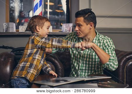 Young Father And His Stylish Little Son In The Barbershop In The Waiting Room.