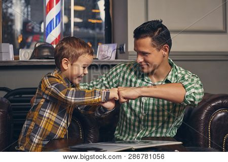 Young Father And His Stylish Little Son In The Barbershop In The Waiting Room.