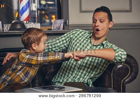 Young Father And His Stylish Little Son In The Barbershop In The Waiting Room.