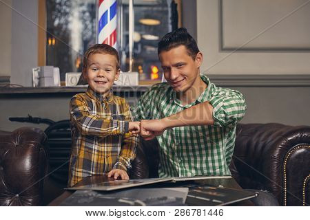 Young Father And His Stylish Little Son In The Barbershop In The Waiting Room.