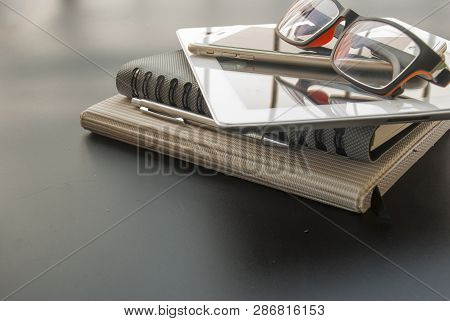 Camera, Glasses, Mobile Phones, Car Keys, Pens, And 2 Notebooks, Placed On A Black Table