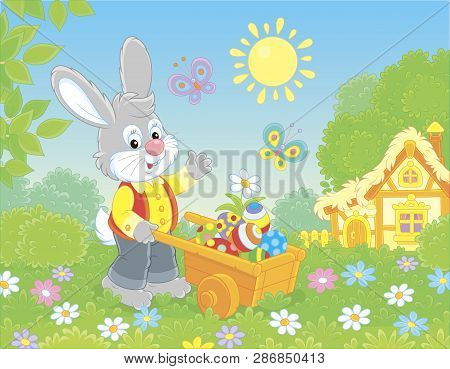 Grey Bunny With A Wooden Handcart Filled With Colorfully Painted Easter Eggs On A Lawn In Front Of A