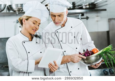 Female And Male Chefs In Iniforms Using Digital Tablet During Cooking In Restaurant Kitchen