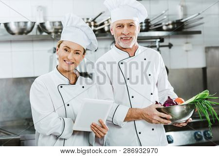 Female And Male Chefs In Iniforms Looking At Camera And Using Digital Tablet While Cooking In Restau