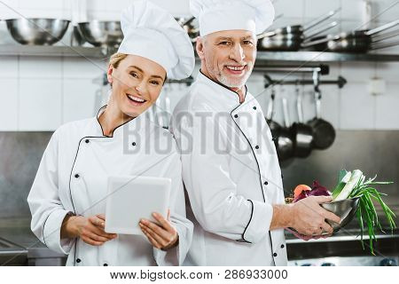 Female And Male Chefs In Iniforms Looking At Camera And Using Digital Tablet While Cooking In Restau