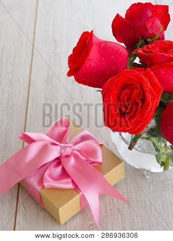 Preparing A Gift, On A Wooden Table, With Roses. Female Hand Holding A Gift, Wrapped With Ribbon, On