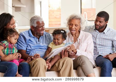 Multi-Generation Family Sitting On Sofa At Home Reading Book With Baby Granddaughters