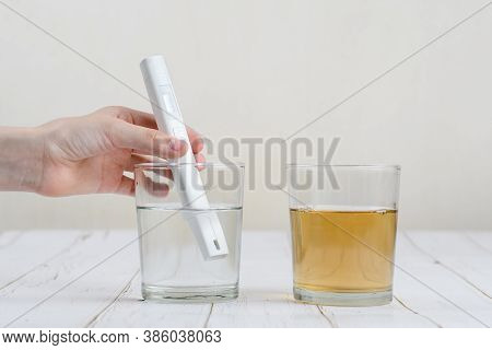 A Hand With A Device For Testing Water Makes A Measurement In A Glass With Transparent Water. Nearby