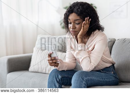 Boredom. Sad Black Lady Spending Time With Smartphone At Home, Sitting On Couch In Living Room, Brow