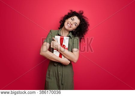 Cute Brunette Woman Embracing A Present Box Smiling At Camera On A Red Studio Wall