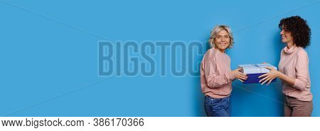 Caucasian Blonde Lady Giving A Present To Her Brunette Curly Haired Friend Posing On A Blue Wall Wit