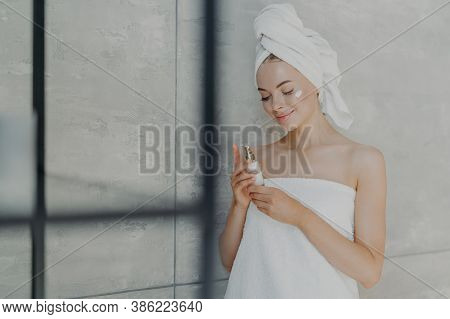 Satisfied Young Caucasian Woman Cares Of Her Beauty, Applies Lotion Cream On Face, Stands Wrapped In