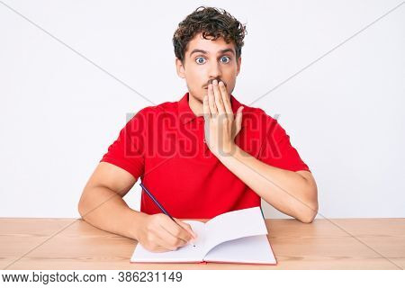 Young caucasian man with curly hair writing notebook sitting on the table covering mouth with hand, shocked and afraid for mistake. surprised expression 