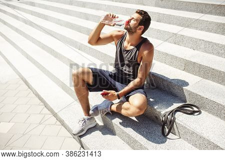 Young Man Exercising Outside. Athlete Sit On Steps Outside And Thursting. Drinking Water And Enjoy I