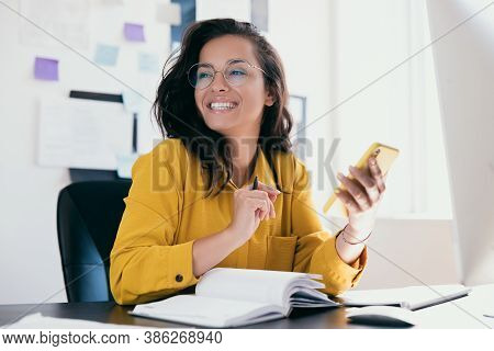 Pretty Cheerful Businesswoman Looking Away Sitting At Desk And Holding Phone In Hand. Positive Think
