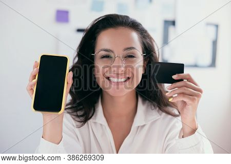 Causasian Cheerful Young Woman With Brown Hair Holding A Card And Smartphone, Smiling And Looking At