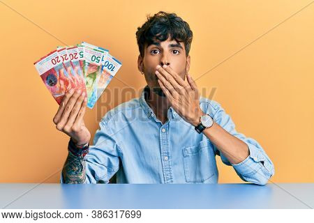 Young hispanic man holding swiss franc banknotes sitting on the table covering mouth with hand, shocked and afraid for mistake. surprised expression 