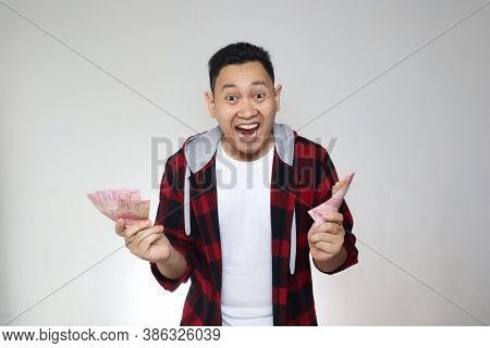 Portrait Of Happy Young Indonesian Man Holding Rupiah Money, Smiling Laughing Winning Gesture, Over 