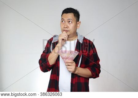 Confused Stressed Asian Man Holding Indonesian Rupiah Cash In Hand, Thinking Wondering How To Spend.