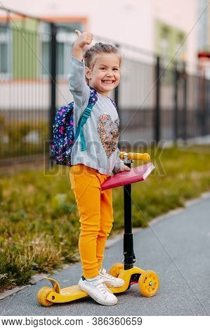 Little Girl Goes To School For The First Time. Little Student With Purple Backpack And Pink Notebook