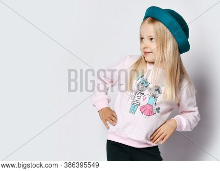 Portrait Of Blonde Kid Girl In Hat, Grey Sweatshirt Sweater And Blue Pants Standing With Hands On Wa