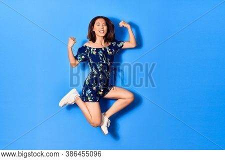 Young beautiful chinese woman wearing casual clothes smiling happy. Jumping with smile on face doing strong geture over isolated blue background