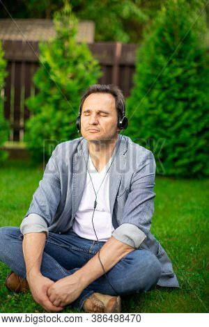 Calm Handsome Adult Man In Headphones Sits On Grass Chilling Outdoors, Listening Relaxing Music With