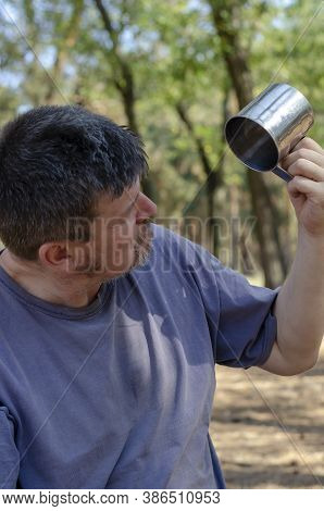 A Man In Bewilderment Looks Into An Empty Metal Mug In The Fores. An Adult Man With Stubble And Gray