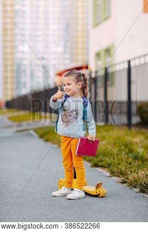 Little Girl Goes To School For The First Time. Little Student With Purple Backpack And Pink Notebook