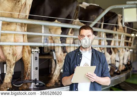 agriculture industry, farming, people, milking and animal husbandry concept - young man or farmer with clipboard and cows at rotary parlour system on dairy farm wearing mask for protection from virus