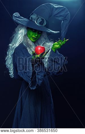 Halloween. Scary green old witch holds out a red apple. Woodland background, evil black magic.