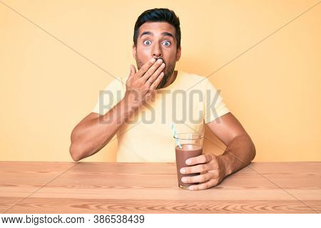 Young hispanic man sitting at the table drinking chocolate beverage covering mouth with hand, shocked and afraid for mistake. surprised expression 