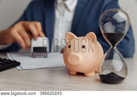 Businessman Approves Contract In Office. Pink Piggy Bank With Stack Of Coins And Hourglass On Wooden