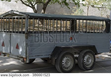 Two-axle Car Trailer On The Roadside. Empty Metal Trailer With A Roof In The Shade Of Trees On A Cit