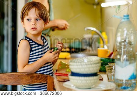 Cute Little Preschooler Boy In Striped T-shirt Help His Mother At Kitchen In Home. Clever Child Cut 