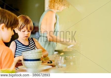 Cute Little Preschooler Boy In Striped T-shirt Help His Mother At Kitchen In Home. Clever Child Cut 