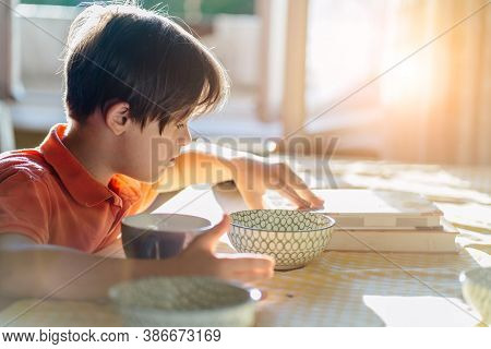 Thoughtful Child Boy Wearing An Orange T-shirt With Down Syndrome Sitting At Table And Reading Book 