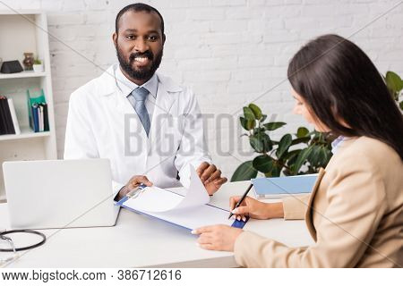 African American Doctor Holding Clipboard With Blank Paper Near Brunette Woman With Pen