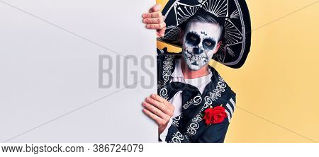 Young man wearing mexican day of the dead costume holding blank empty banner looking positive and happy standing and smiling with a confident smile showing teeth 