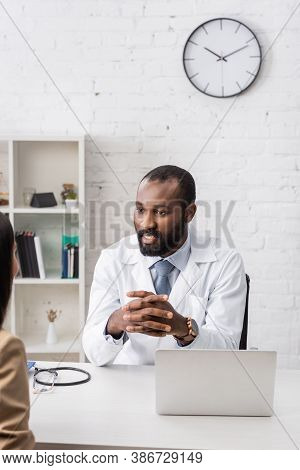 Selective Focus Of African American Doctor Sitting With Clenched Hands Opposite Patient At Workplace